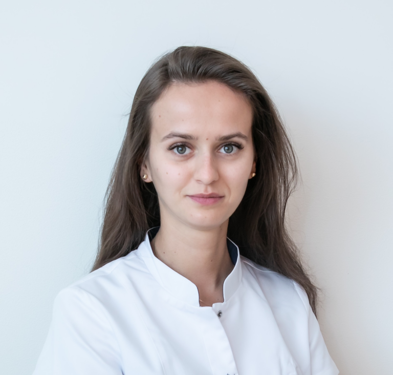 Marina Nieborak, młoda kobieta o długich brązowych włosach, ubrana w biały fartuch lekarski, stoi na jednolitym, białym tle, patrząc prosto w kamerę z neutralnym wyrazem twarzy. niebodent bemowo