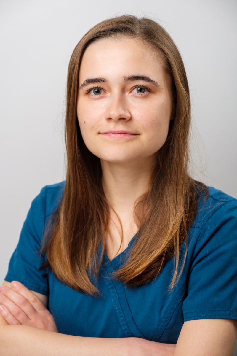 Marina Nieborak, młoda kobieta o prostych brązowych włosach i spokojnym wyrazie twarzy, stoi ze skrzyżowanymi ramionami, ubrana w turkusowo-niebieski fartuch medyczny na jednolitym, jasnym tle. niebodent bemowo