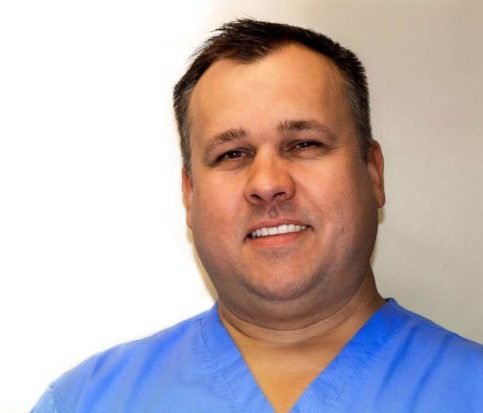 Mężczyzna o kr&oacute;tkich, ciemnych włosach, ubrany w niebieski fartuch medyczny, stoi uśmiechnięty na jasnym tle. Sfotografowała go Marina Nieborak. niebodent bemowo