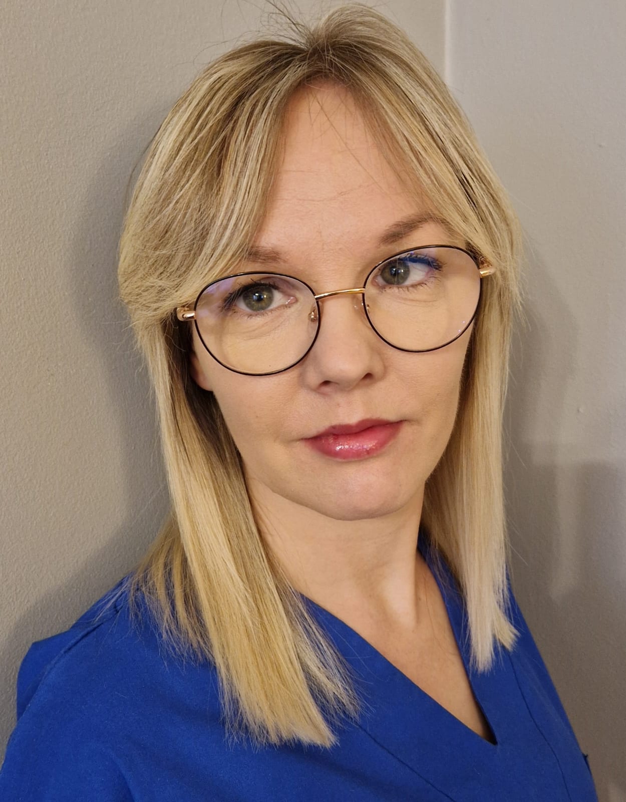 Blondynka w okularach i niebieskim stroju medycznym, patrząca w kamerę. Portret przedstawiciela służby zdrowia.